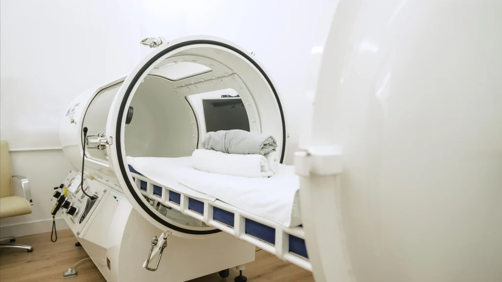 Open hyperbaric oxygen therapy chamber with white bedding and control panels inside a clinical room